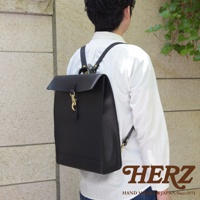 黑色皮革紙袋風後背包，配有金色金屬扣環和皮革背帶，由男性背於身後，身穿白色襯衫。