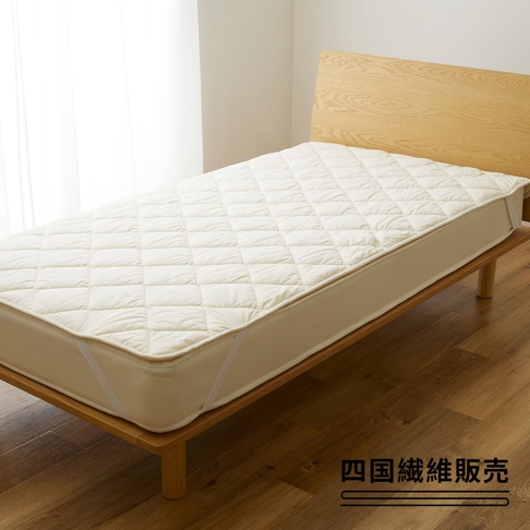 米白色菱格紋鋪棉床墊，邊緣有鬆緊帶，放置於木製床架上，背景為淺色牆壁與白色窗簾。