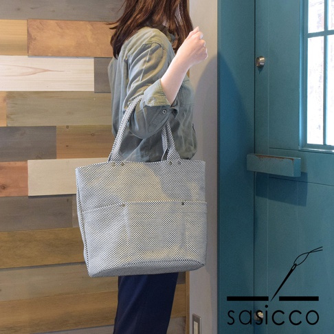 一位女性身穿綠色襯衫，肩揹一個白色和黑色棋盤格圖案的三河木綿刺子織大開口托特包，包包有兩個外部口袋，並以鉚釘固定。