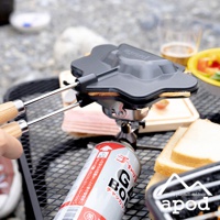 黑色熱壓三明治烤夾，配有木質手柄，夾著一片三明治，放置於瓦斯罐上方，周圍有吐司、火腿和配料。