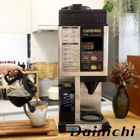 銀色與黑色系的自動生豆烘焙咖啡機 MC-520A，配有液晶顯示螢幕、控制面板及咖啡壺，旁邊有一個裝有乾燥植物的深綠色花瓶。