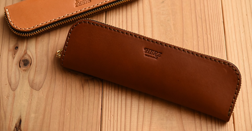 職人手作皮革托盤式收納筆袋KP-5，深棕色皮革，配金色拉鍊，上面壓印有HERZ品牌標誌。