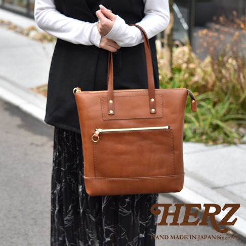 一位女性手提著日本HERZ職人手作的棕色皮革托特包，包款採用簡約的直式設計，正面設有拉鍊外袋與金屬拉環，包身以厚實的皮革製作，整體呈現出精緻的工藝質感，右下方印有品牌標誌。