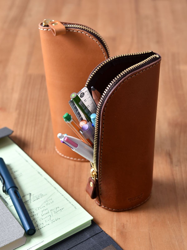 兩個棕色皮革製成的直立式收納筆袋，其中一個筆袋拉鍊打開，露出內部的筆、尺等文具，另一個筆袋拉鍊閉合。