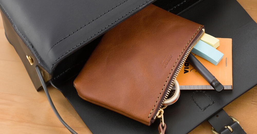 一個棕色皮革長方形拉鍊收納包，拉鍊部分露出，內有筆、筆記本和便利貼，收納包放在一個黑色皮革包內。