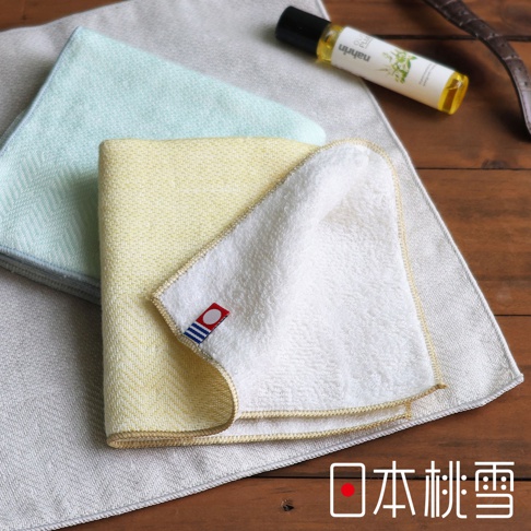 日本桃雪今治蓬鬆人字紋紗布方巾，含淺綠色、黃色與白色毛巾，上有一小小日式旗幟標籤，背景為木紋桌面，右側有一小瓶精油，圖片下方有日文「日本桃雪」字樣。