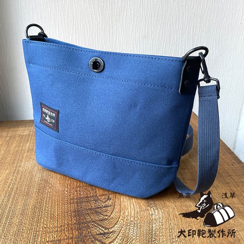 犬印鞄製作所的藍色純綿帆布迷你側背包，帶有品牌標籤和金屬釦，放置於木質桌面上，右下角有品牌標誌。