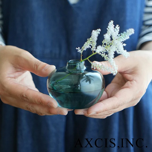 AXCIS, INC. 藍綠色玻璃小花瓶，兩隻手捧著，瓶中插著一束白色細緻的花。
