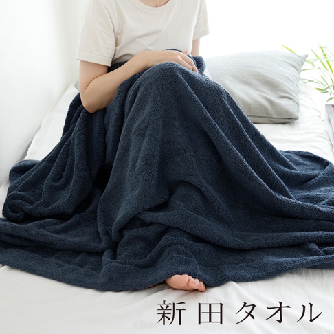 一位身穿白色短袖上衣的女性坐在白色床鋪上，身上覆蓋著一條厚實柔軟的深藍色長毛絨毯子，毯子垂墜感自然並蓋住雙腿，露出腳趾，圖片下方標註新田タオル文字，整體呈現居家舒適與溫馨的氛圍。