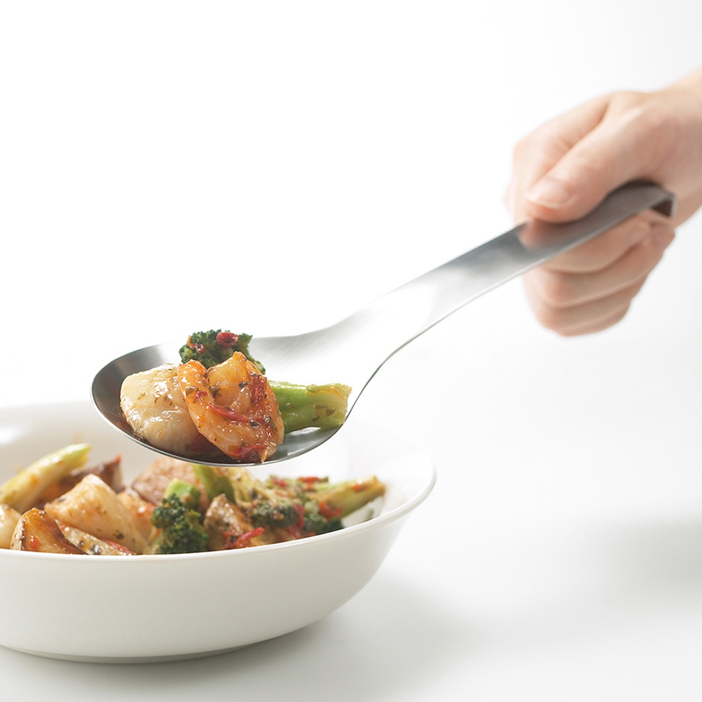 一只手正握著一把銀色不鏽鋼服務餐匙，餐匙上盛著鮮蝦、干貝、花椰菜和烤馬鈴薯塊，準備舀起碗中同樣的混合菜餚。