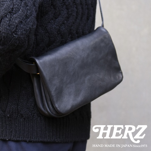 黑色皮革斜背包，帶有翻蓋和金色金屬扣，斜挎於穿著黑色針織衫的人身上。