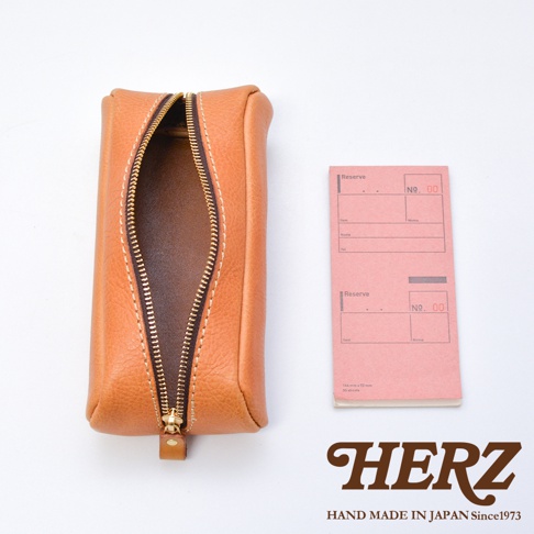 一個日本 HERZ 手工職人製作的棕色皮革收納筆袋，採用優質柔軟皮革製成，表面具有細緻紋理，筆袋呈圓弧長型，頂部設有金色金屬拉鍊，拉鍊開口處呈現自然的弧度，袋身側邊附有一個小型皮革拉環。筆袋旁放置了一本粉紅色的直式橫線記事本，整體風格簡約且具有溫潤的手作質感。