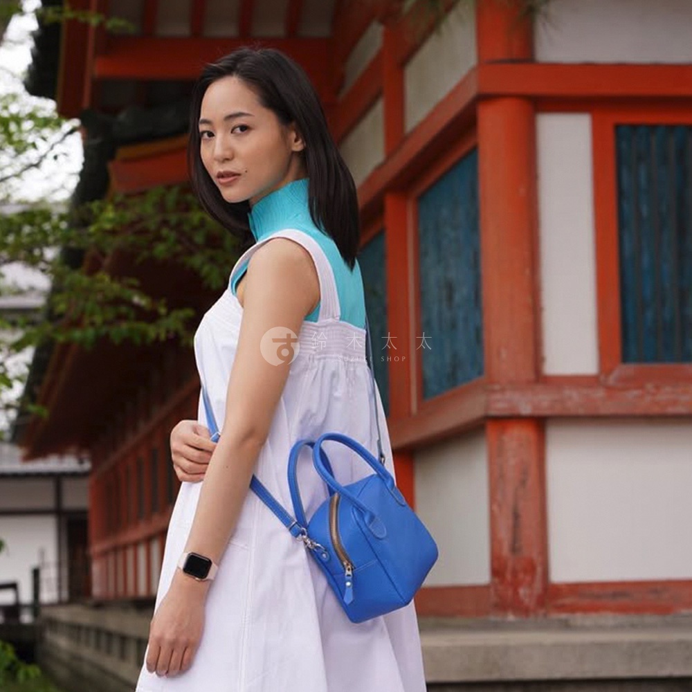 身著白色洋裝的女子，斜肩背著一個亮藍色、造型類似貝殼的皮革手提包，包包有兩個提把和一個可拆卸肩帶，並配有金色拉鍊。