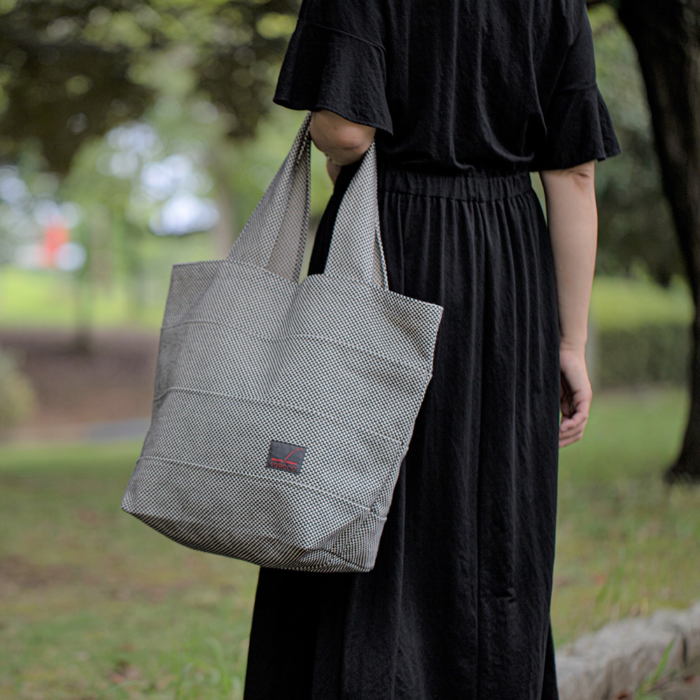 一手提著灰白色細格紋刺子織大容量托特包，包上有紅色品牌標籤，搭配黑色長裙，置於戶外綠色背景前。