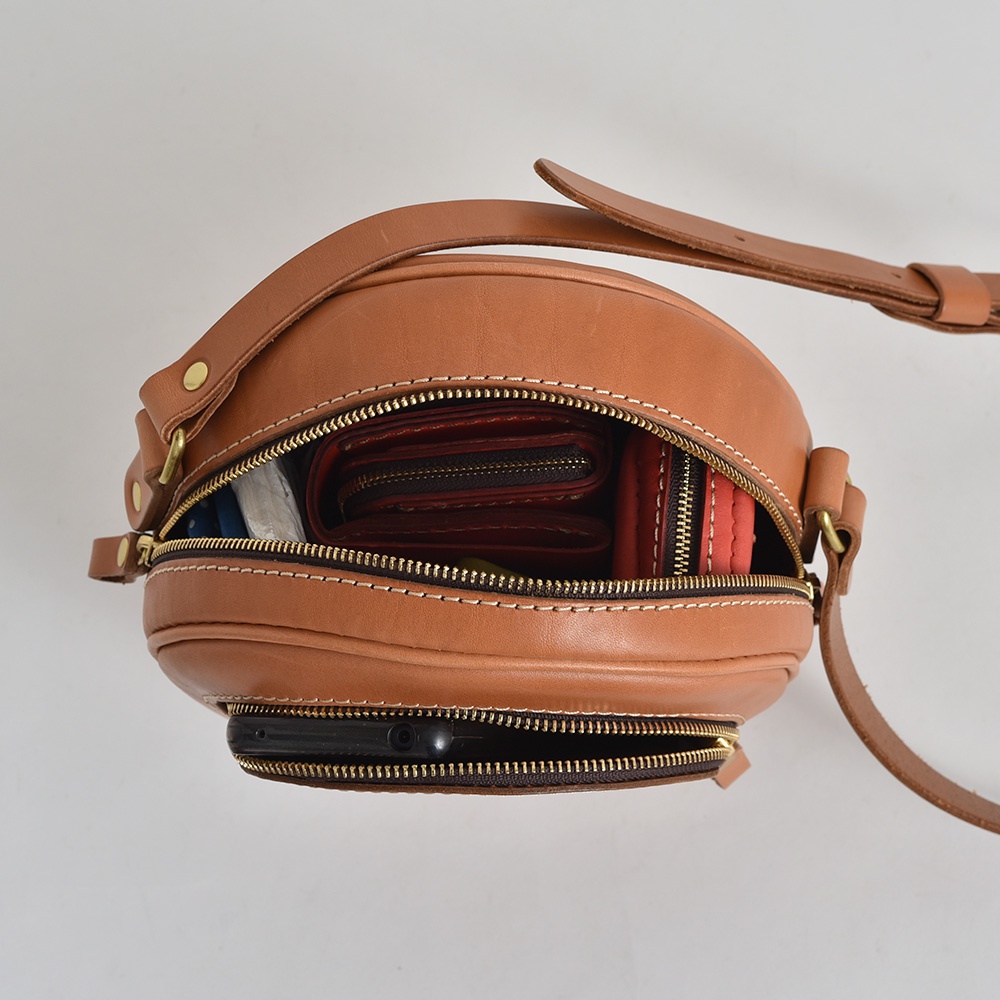 職人手作皮革小圓包，棕色皮革材質，圓潤造型，頂部拉鍊開口露出內部的紅色錢包和白色物品，底部拉鍊開口可見手機。