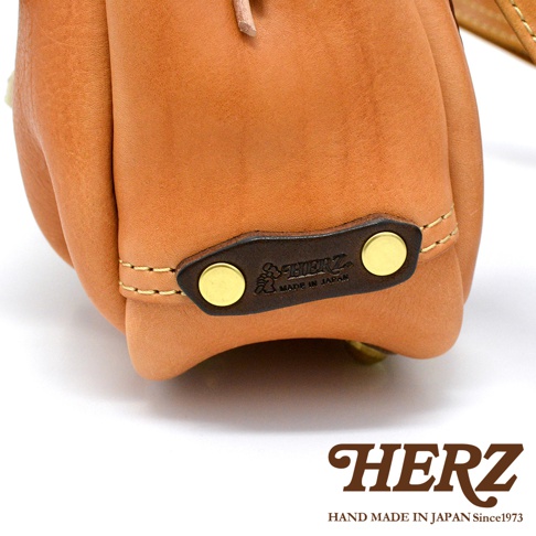 淺棕色皮革郵差包，特寫鹿角扣細節，深棕色皮革裝飾片上壓印有「HERZ」品牌標誌及「MADE IN JAPAN」字樣，以金色卯釘固定，縫線為淺黃色。