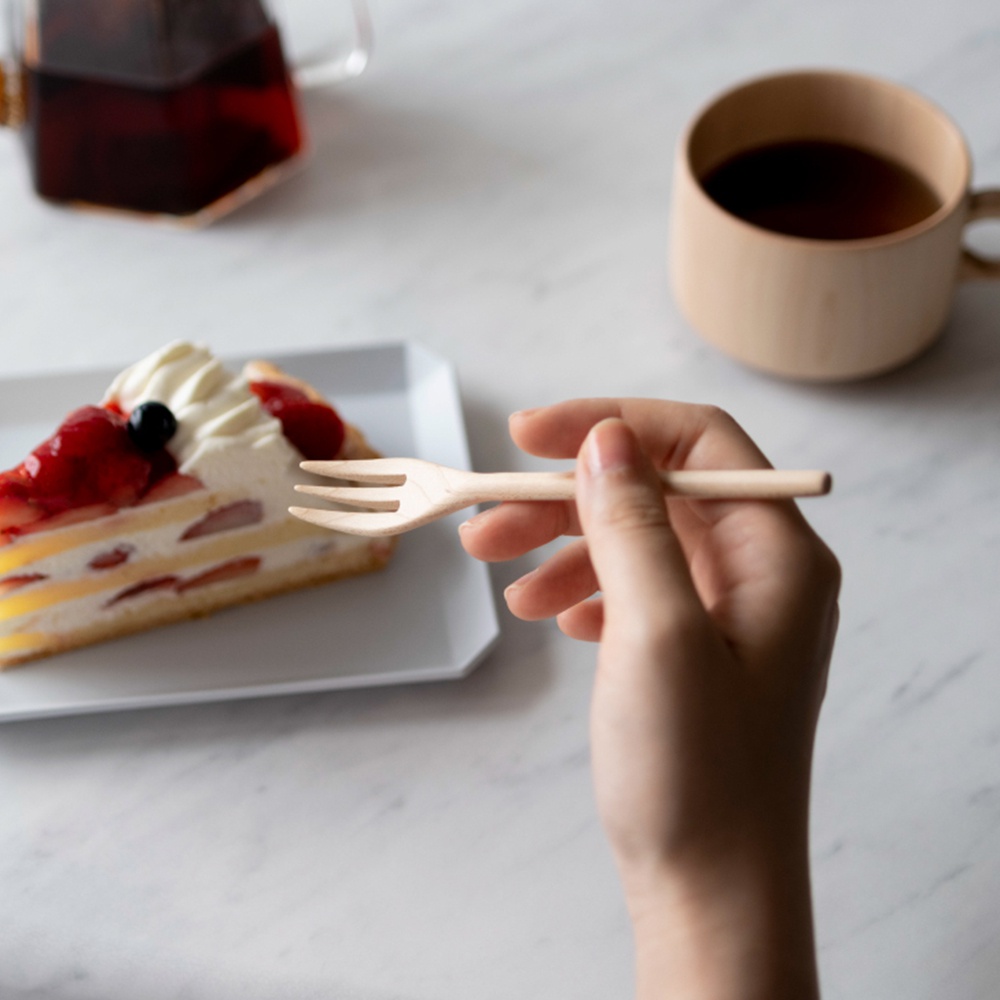一隻手拿著楓木點心叉，準備享用盤中的草莓鮮奶油蛋糕。背景是淺色的木紋桌面，一旁還有一個木質咖啡杯。
