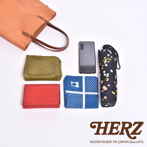 頂視圖展示一個日本HERZ手工皮革托特包及其收納物品，包含一個淺棕色皮革托特包、橄欖綠色皮革錢包、紅色皮革皮夾、一台黑色Sony手機、一條藍白波點縫紉圖案手帕，以及一把黑色花卉圖樣折疊傘，右下角印有HERZ品牌標誌與文字。