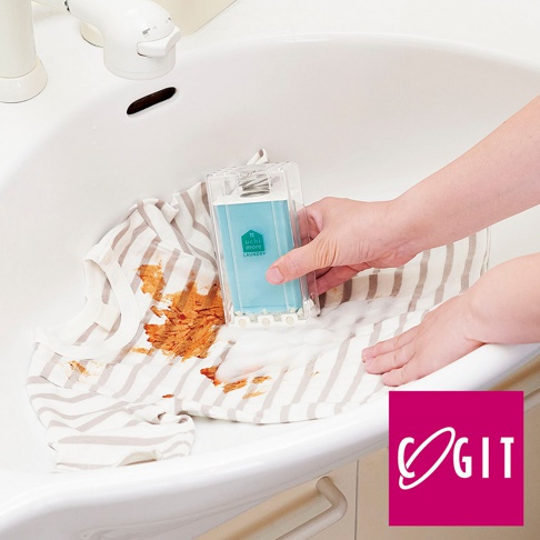 一手持透明塑膠盒裝的淺藍色洗衣皂刷，對著沾有咖啡漬的條紋衣物進行局部清洗。