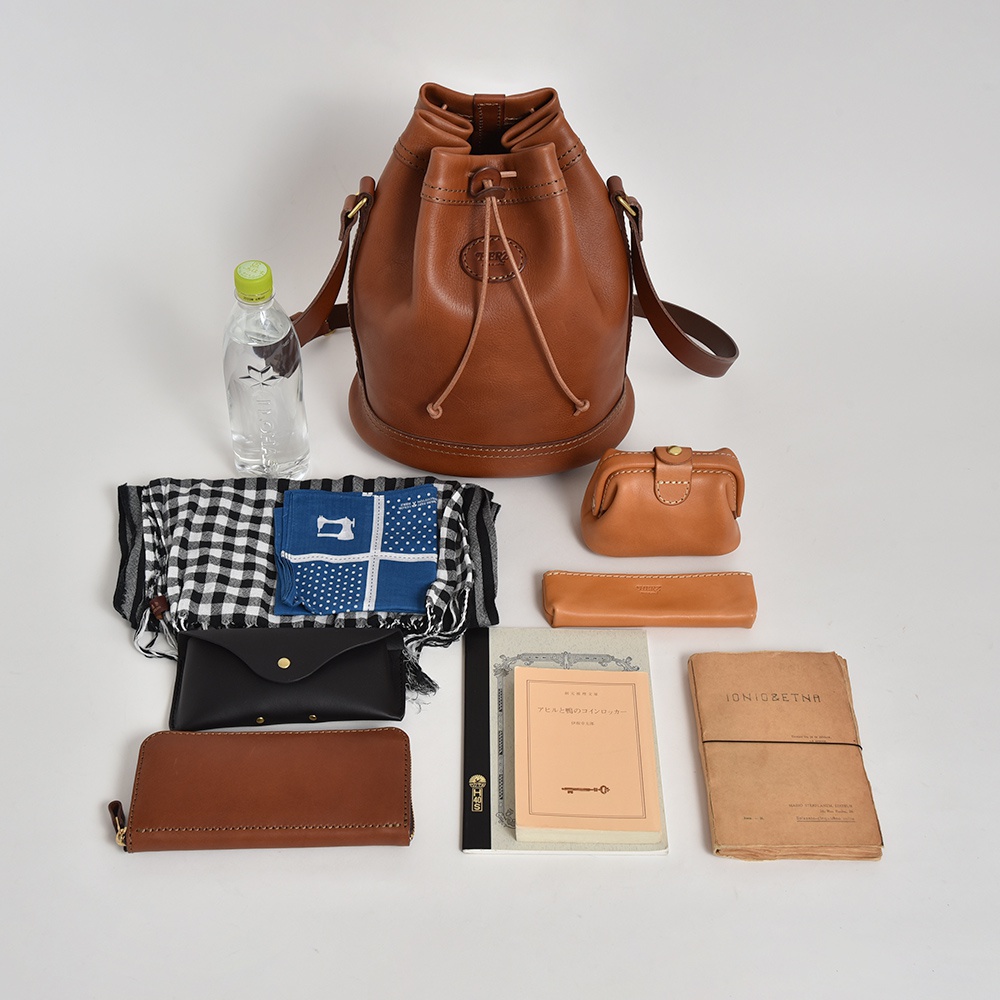 職人手作皮革束口單肩包，呈現經典棕褐色，搭配同色系皮革束繩與品牌標誌。周邊配件包含一瓶礦泉水、黑白格紋圍巾、藍色圖案手帕、黑色皮夾、棕褐色長夾、小化妝包、筆記本及一本泛黃的書本。