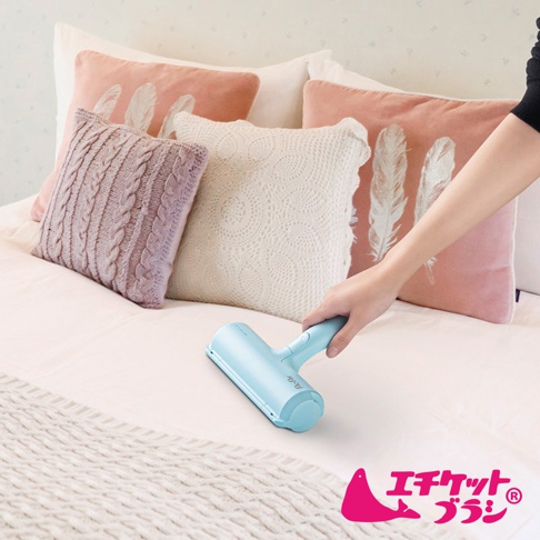 淺藍色免耗材抗菌棉被除塵刷，置於床鋪上，旁邊有粉色、紫色和白色靠墊，其中粉色靠墊印有白色羽毛圖案，白色靠墊為蕾絲編織。