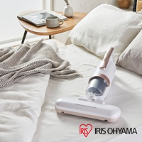 白色與玫瑰金色的手持式無線吸塵器，配有透明集塵筒和寬大的吸頭，放置於鋪有白色床單的床上，旁有棕色菱格紋毯子及茶几。