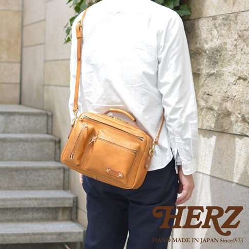 皮革兩用多功能收納單肩包，採用天然皮革製成，呈現溫潤的淺棕色。包款設計有多個拉鍊口袋和一個前置小口袋，方便收納，適合斜背或肩背。