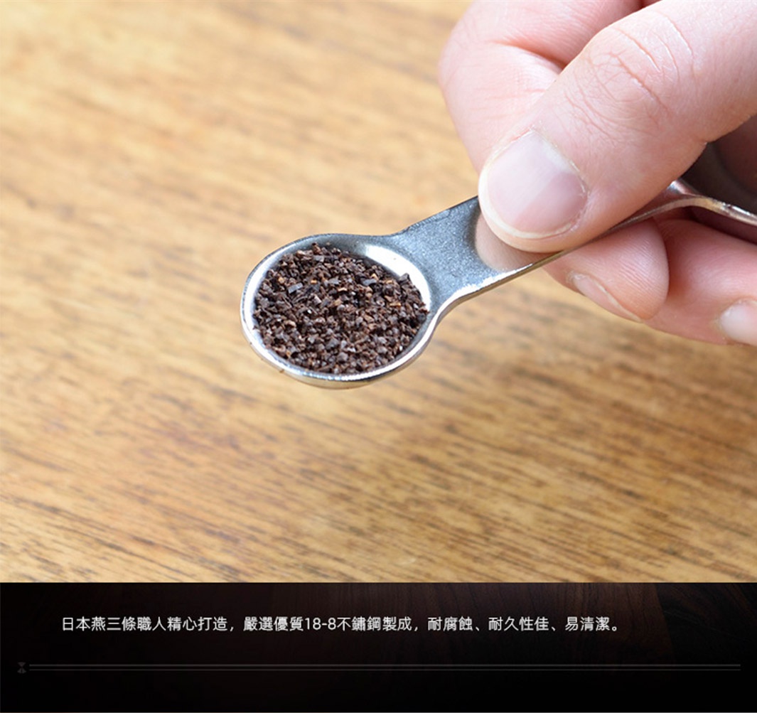 一只手拿著一個日製18-8不鏽鋼雙頭計量式咖啡豆勺/量匙，量匙中裝有咖啡豆渣，勺子呈現銀色金屬光澤，背景是木紋桌面。