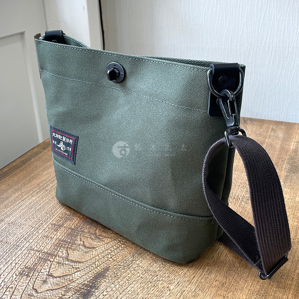 一個綠色帆布迷你側背包，帶有黑色肩帶和金屬扣。包包上有一個紅色標籤，上面印有日文文字。
