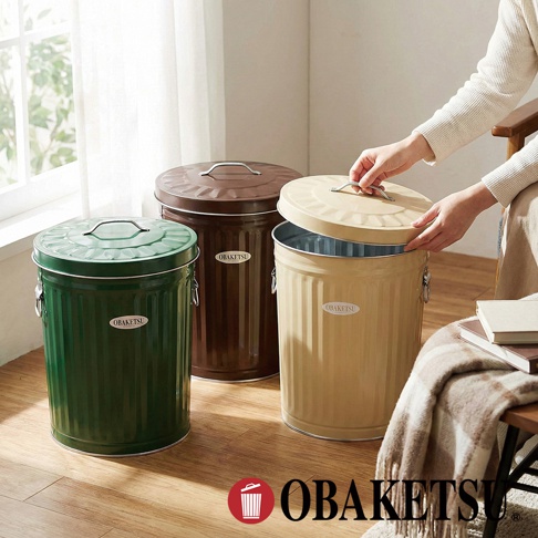 OBAKETSU 品牌手工製金屬垃圾桶，共有綠色、棕色和米黃色三款，其中米黃色垃圾桶的蓋子由手部提起，展示了其經典的圓筒造型和壓紋設計。