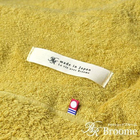 細節特寫：一條黃綠色、觸感柔軟的今治毛巾被，上面縫有品牌標籤，標籤上印有「made in Japan」和「La vie avec Broome」字樣，以及一個小巧的紅白色圓點和藍色條紋的裝飾標識。