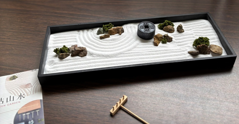 一個長方形黑色木質底座的日本枯山水模型，沙盤內細緻的白色沙粒上繪有圓形波紋，搭配數顆天然岩石、微縮青苔與一個刻有漢字的黑色圓形擺件，前方放有一支木製耙子，左側則有印著枯山水名稱的展示卡。