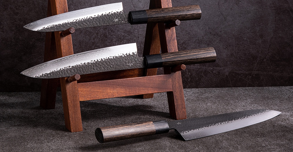 三把「玄 / Utility knife 多功能刀16cm」，刀身為銀色金屬，帶有錘紋和 Damascus 紋理，刀柄為深棕色木質，配有黑色金屬連接環，置於木質刀架上。
