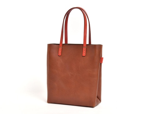 職人手作皮革直式軟皮托特包，深棕色皮革主體，搭配亮橘色皮革提把，提把與包身以白色縫線連接，包身側面有一個紅色小標籤。
