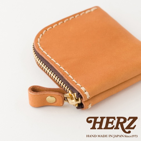 這是一張日本HERZ職人手作皮革零錢包的特寫照片，展示了淺棕色皮革材質與細緻的縫線工藝，配有金色拉鍊及帶有鉚釘扣的皮革拉環，右下角印有HERZ品牌商標與產地資訊。
