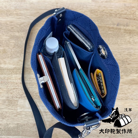 深藍色刺子織迷你側背包，內部整齊擺放著手機、錢包、筆記本、水瓶及鑰匙包，材質為紋理感的織布，搭配銀色金屬釦。