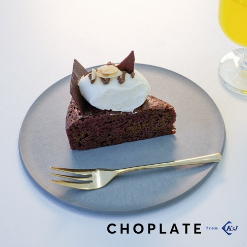 灰色的 CHOPLATE 兩用砧板盤上擺放著一塊巧克力蛋糕，蛋糕頂部覆蓋著白色鮮奶油、杏仁片和巧克力薄片，盤旁有一支金色的叉子。