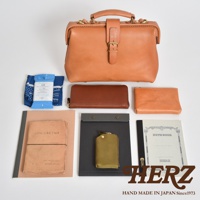 一組職人手作皮革配件，包含一個大型的駝色手提包，側邊有金屬扣環，包身配有皮帶和金屬扣。另有兩個長夾，一個為深棕色，一個為淺棕色。配件包含藍色印花布、藍色濕紙巾包、粉紅色筆記本、黑色文件夾、綠色鑰匙包以及一本淺灰色的筆記本。