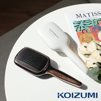 白色和黑色兩款負離子養髮梳，黑色款帶有玫瑰金飾邊，放置於白色桌面上，背景有雜誌和花朵裝飾