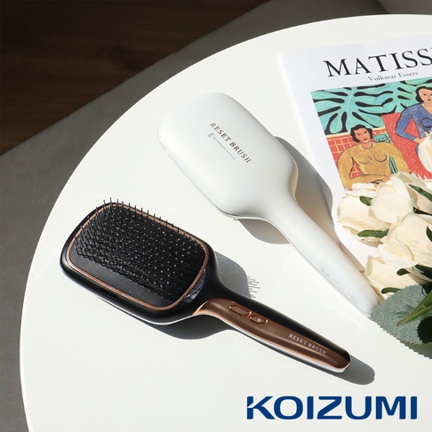 白色和黑色兩款負離子養髮梳，黑色款帶有玫瑰金飾邊，放置於白色桌面上，背景有雜誌和花朵裝飾