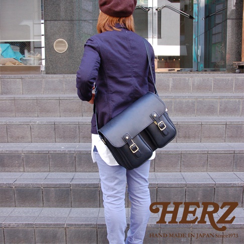日本HERZ職人手作皮革經典雙扣郵差包，側背於身穿海軍藍外套的模特兒身上。