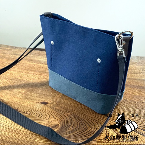 犬印純綿帆布迷你側背包，採用深藍色帆布與淺灰色帆布拼接設計，配有黑色織帶和銀色金屬扣環。