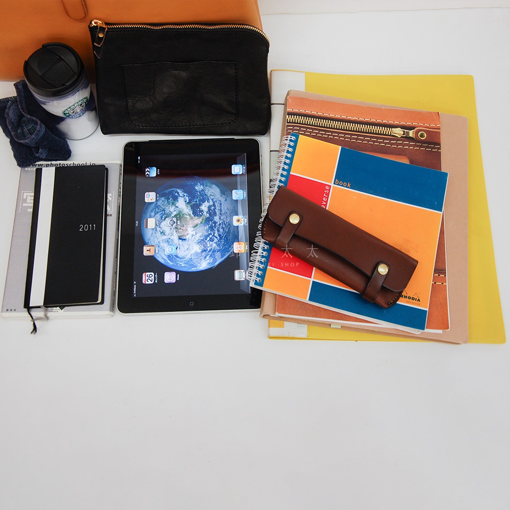 職人手作皮革軟式杜勒斯包，棕色皮革筆套，置於多本雜誌與筆記本上方，一旁有黑色皮夾、咖啡杯及牛仔布料。