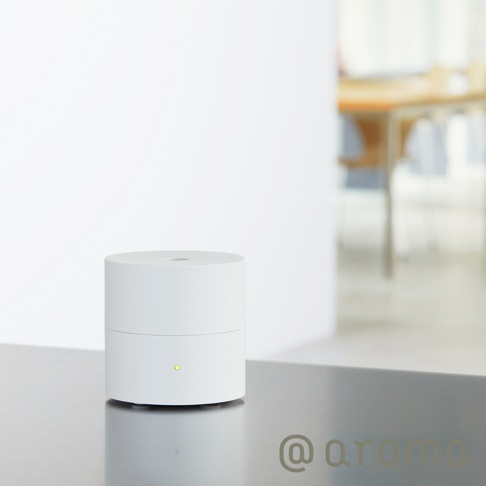 白色圓柱形隨身精油香氛機，頂部有出孔，底部有一個綠色指示燈，置於金屬檯面上，背景為模糊的室內空間。