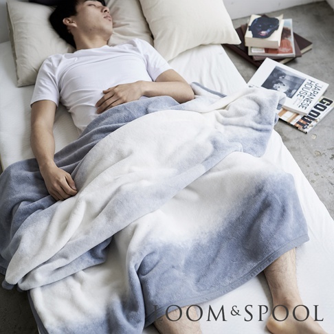 印有「LOOM & SPOOL」標誌的單人短絨毯，採用漸層藍白色調，質地柔軟，蓋在一位正在睡覺的男性身上。