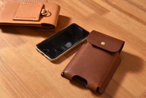 職人手作皮革三用手機收納包，包含一個淺棕色可穿過皮帶的收納包，以及一個深棕色帶有黃銅金屬鈕扣的直立式手機套。旁邊擺放著一部黑色螢幕手機。
