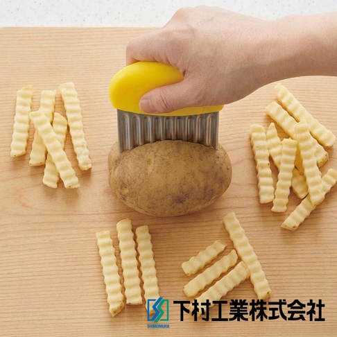 一隻手握著黃色握把的下村工業波浪造型切刀，正在切割木質砧板上的馬鈴薯，周圍擺放著多根已切好的波浪狀薯條，展現波浪切刀的使用情境。