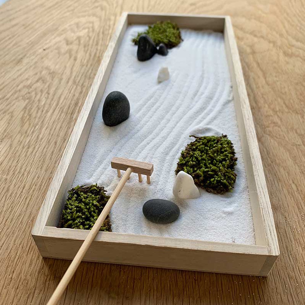 淺木色長方形托盤內，鋪著白色細沙，沙上刻有波浪紋。沙中點綴著綠色苔蘚、黑色和白色鵝卵石，以及一把小型木製耙子。