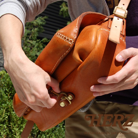 棕褐色皮革郵差包，有著復古金屬扣環和手工縫線，包身柔軟且容量大，展現職人手作質感。