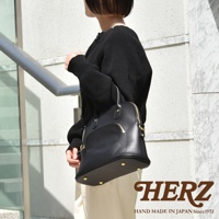 一位女性手持黑色經典貝殼造型皮革手提包，包身光滑，設有金色拉鍊和金屬腳釘。